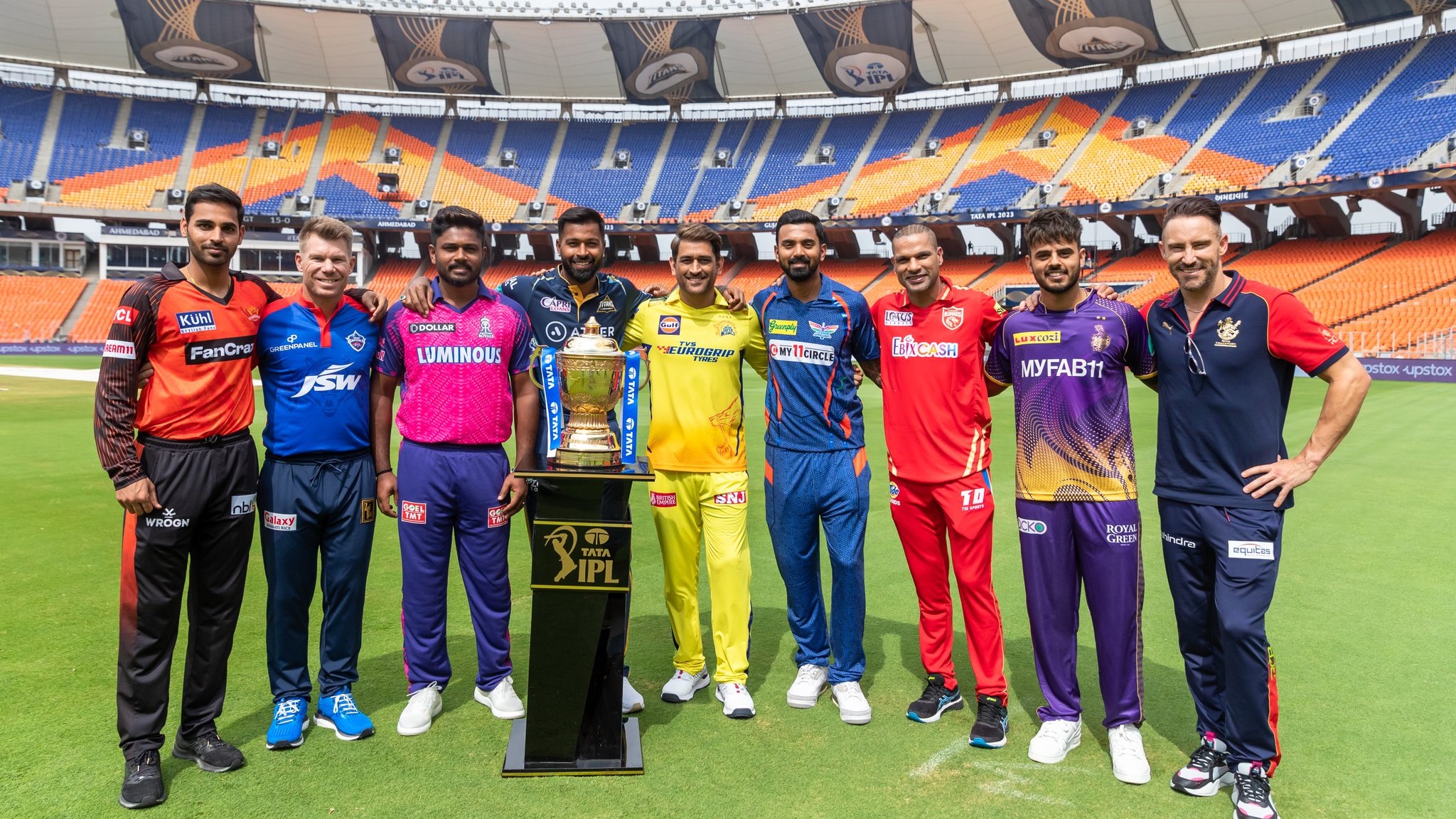 IPL team captains standing around the Indian Premier League trophy in a large cricket stadium, representing different franchises ahead of the tournament season.