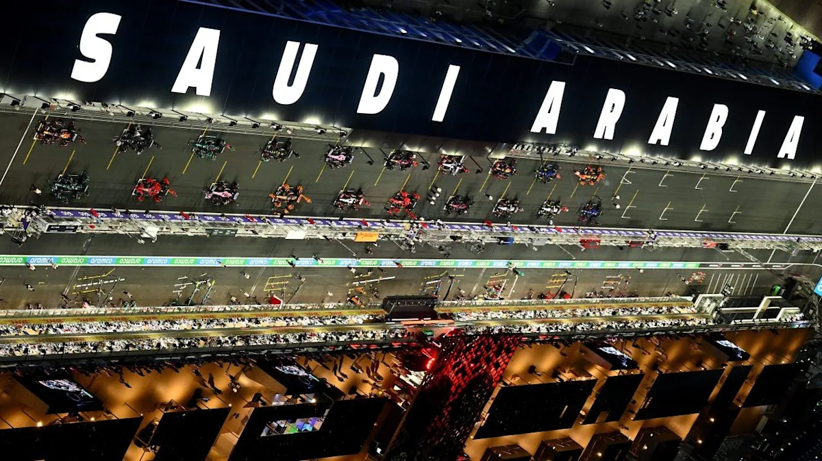 Aerial view of Formula 1 pit lane in Saudi Arabia, featuring race cars, team garages, and illuminated circuit during a night Grand Prix event.