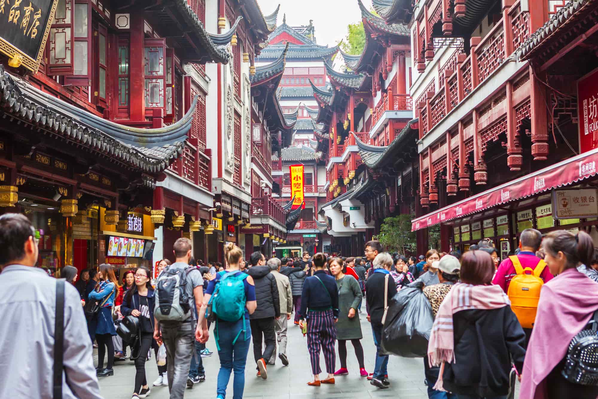 Crowded Shanghai old town street with traditional Chinese architecture, bustling market shops, and tourists exploring historic Yuyuan Bazaar district.