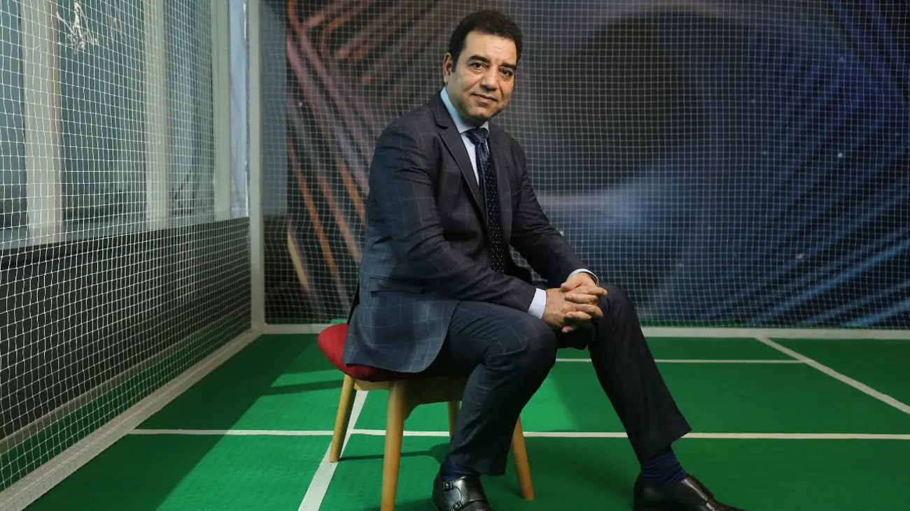 Business executive in a suit seated on a chair inside an indoor sports court, blending corporate leadership with sports or athletic branding context.