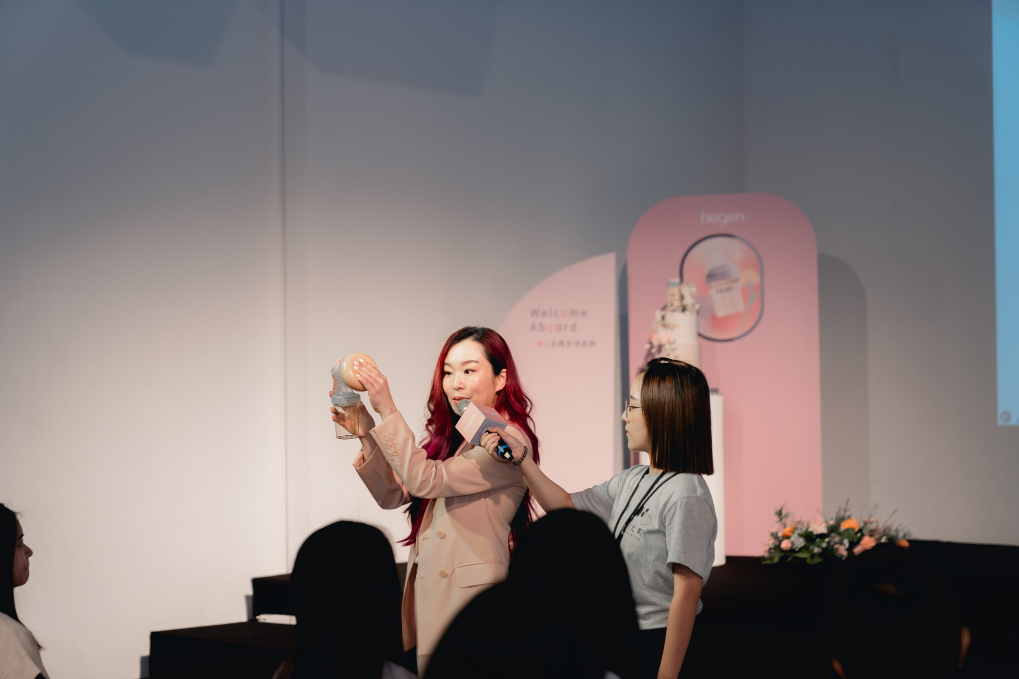 Yvon Bock demonstrating a baby product on stage while holding a microphone, with an assistant beside her.
