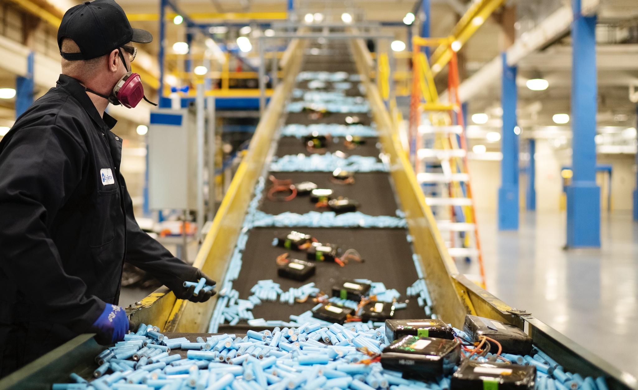 Factory worker inspecting lithium-ion battery cells on a conveyor belt in an industrial recycling or manufacturing facility, highlighting EV battery processing and supply chain operations.