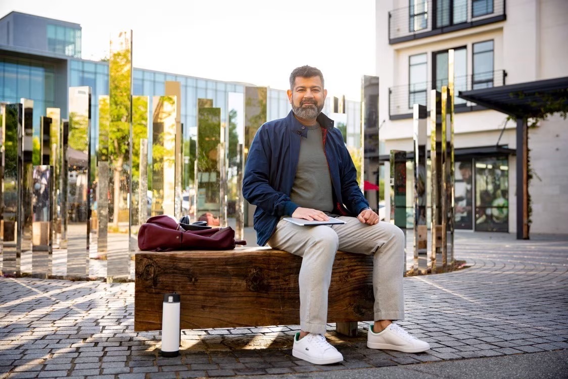 Man sitting on a wooden bench with a tablet in an urban plaza, surrounded by modern architecture and reflective art installations, representing remote work and digital lifestyle.