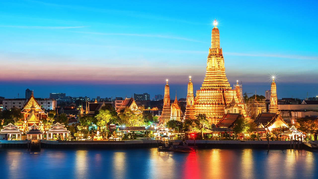 Wat Arun temple illuminated at sunset along the Chao Phraya River in Bangkok, showcasing Thailand’s iconic cultural landmark and vibrant cityscape.