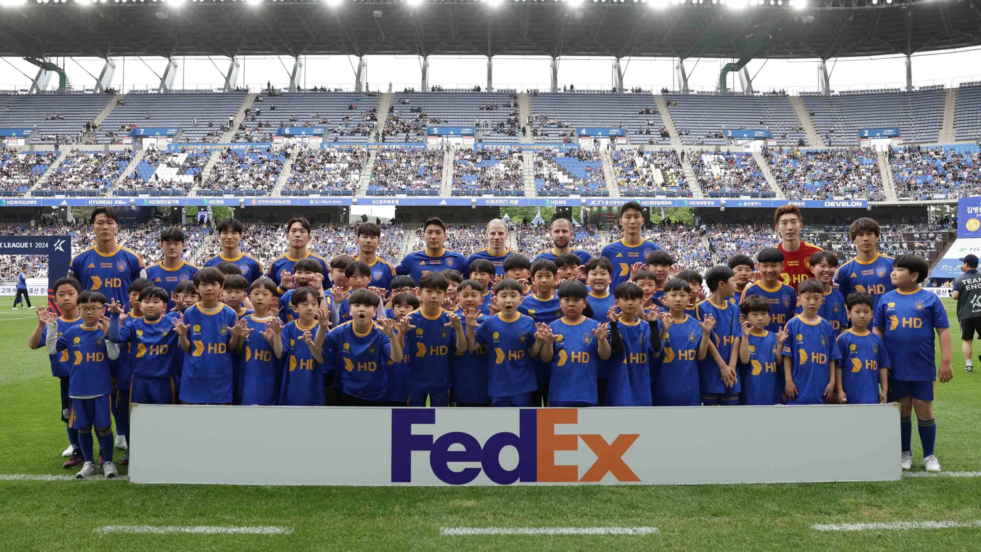 Professional football players and youth mascots posing on the field before a K League 2024 match in South Korea, with packed stadium seating and FedEx sponsorship branding visible.